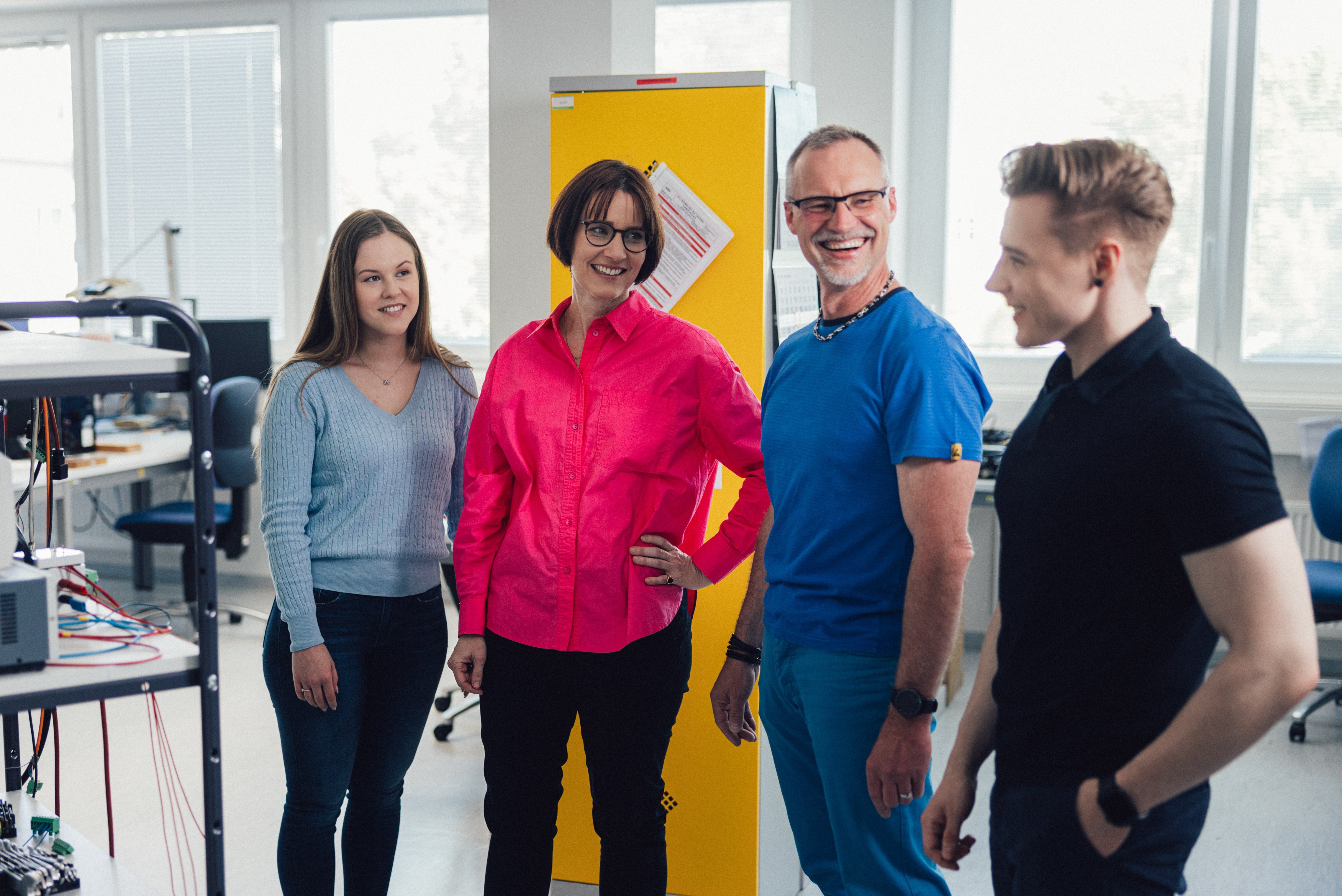 The image shows four employees standing close together in a room with technical equipment, talking and loughing.